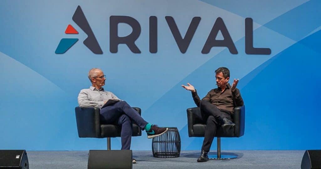 Two people are seated in armchairs on a stage with a large blue background displaying the logo "ARIVAL." The person on the left casually sits with one leg crossed over the other wearing a grey shirt and dark trousers, while the person on the right is gesturing with their hands, dressed in all black. There are speakers on the stage floor in front of them.