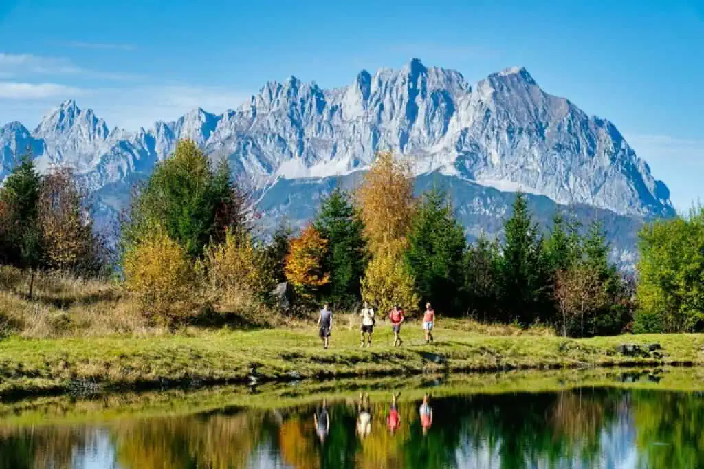 Four people hike along a trail near a reflective lake, their journey embodying experiences in nature, with a backdrop of tall, rocky mountains under a clear blue sky.