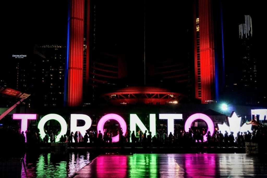 "Toronto" sign lit up at night with crowds gathered in front, reflecting in water