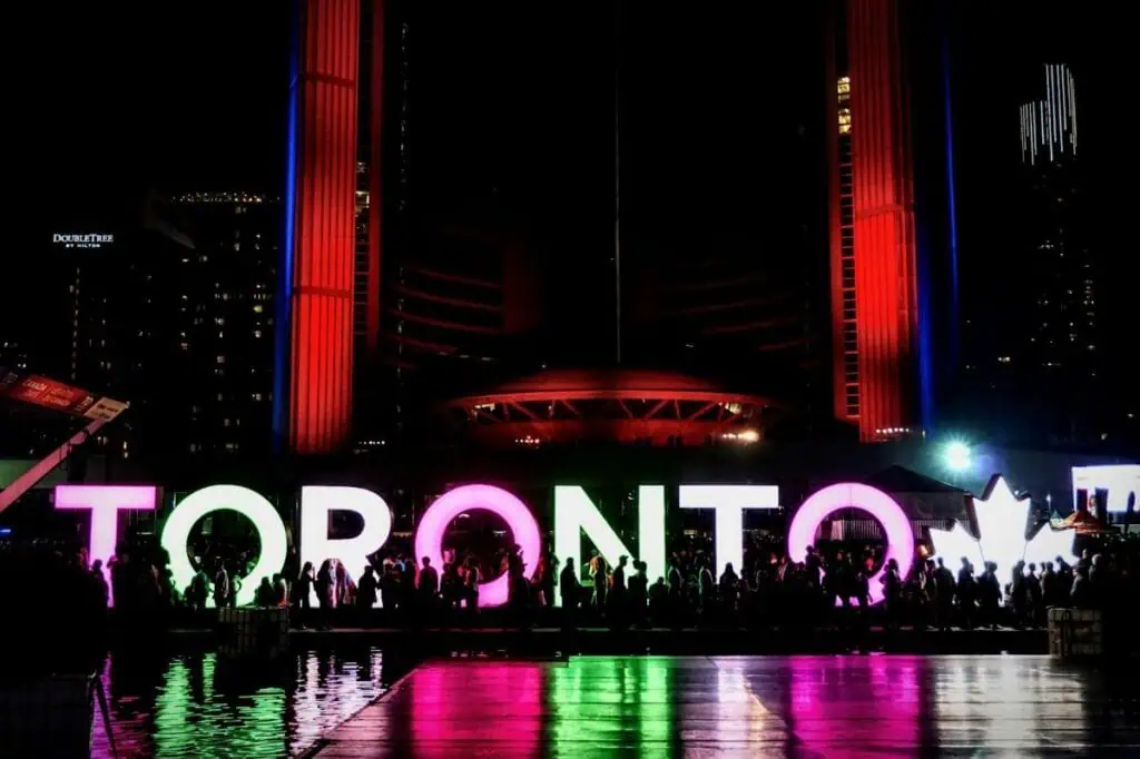 "Toronto" sign lit up at night with crowds gathered in front, reflecting in water