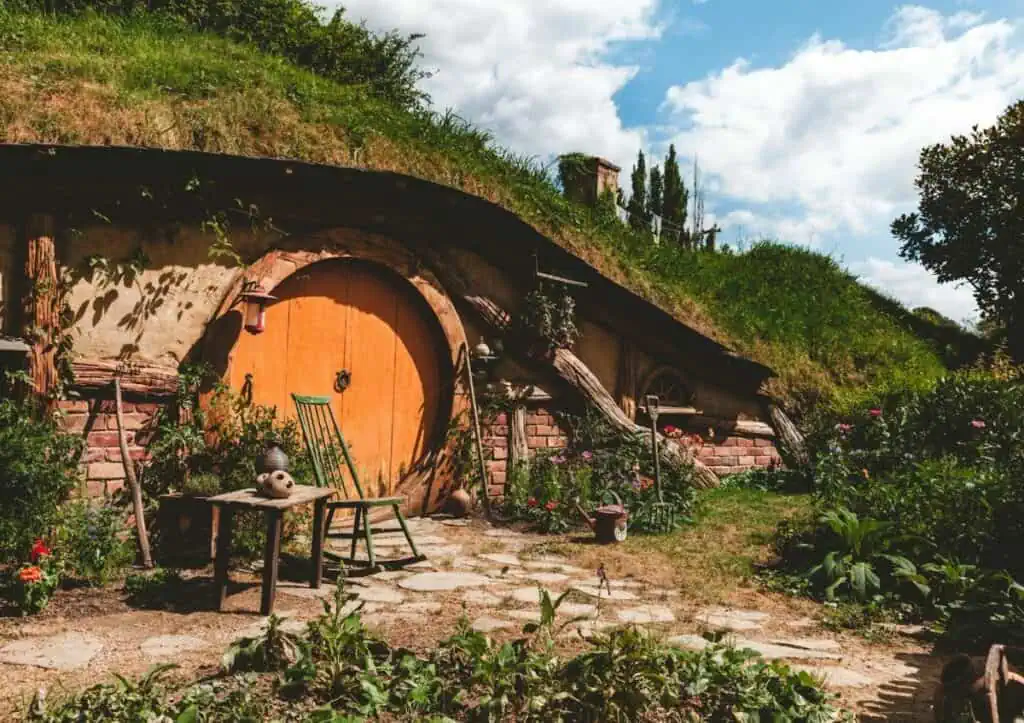 A hobbit house with a round orange door, surrounded by greenery and flowers, with a green chair and small table on the stone patio.