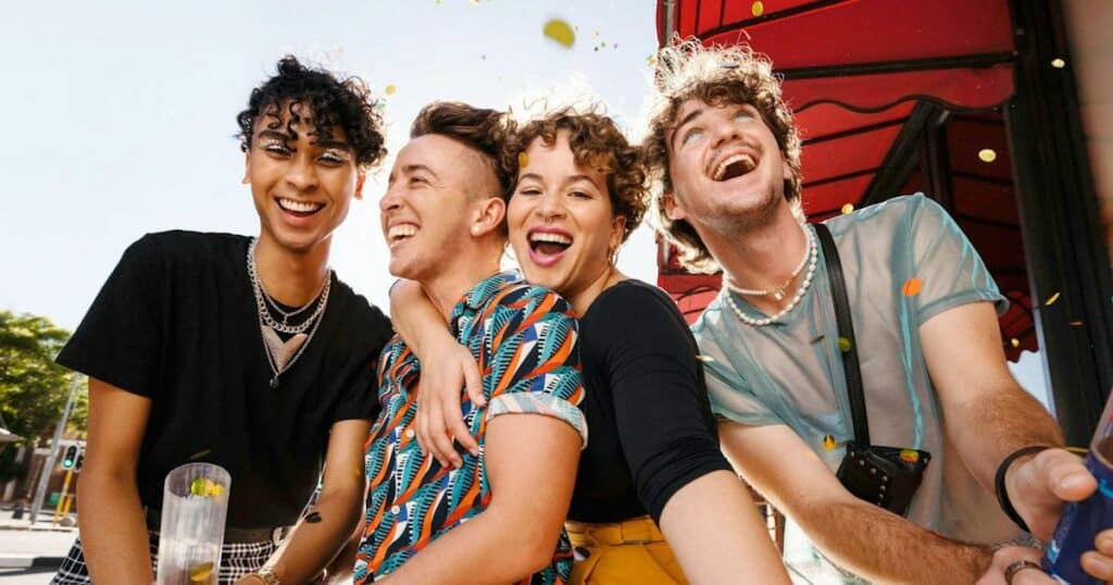 Four young adults laughing and smiling together outdoors. They appear diverse in appearance and style, with colorful clothing and accessories. The image conveys a sense of joy, friendship, and celebration, aligning with the article's focus on LGBTQ+ travelers and their vacation experiences.