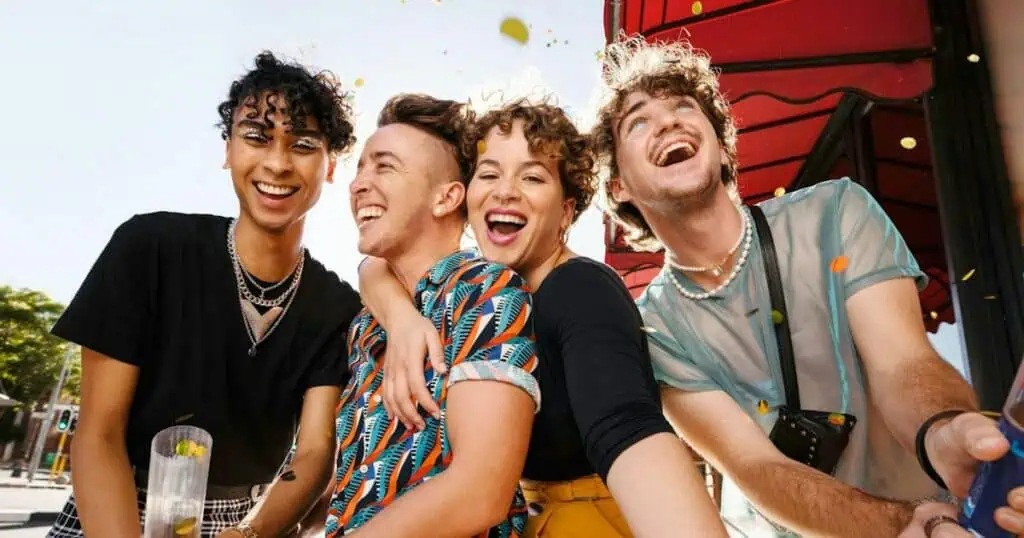 Four young adults laughing and smiling together outdoors. They appear diverse in appearance and style, with colorful clothing and accessories. The image conveys a sense of joy, friendship, and celebration, aligning with the article's focus on LGBTQ+ travelers and their vacation experiences.