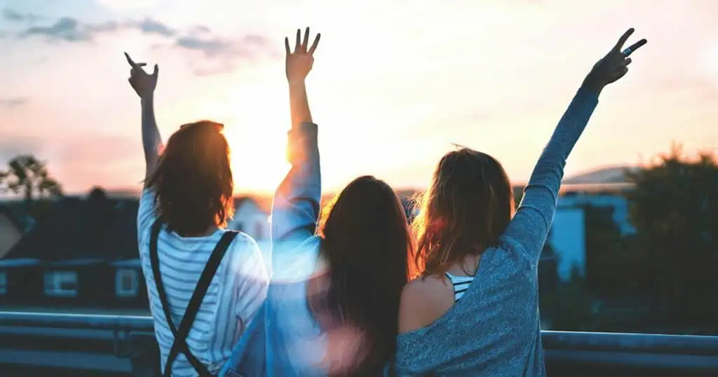 Three friends raise their arms joyfully against a sunset sky, silhouetted in a celebratory moment.